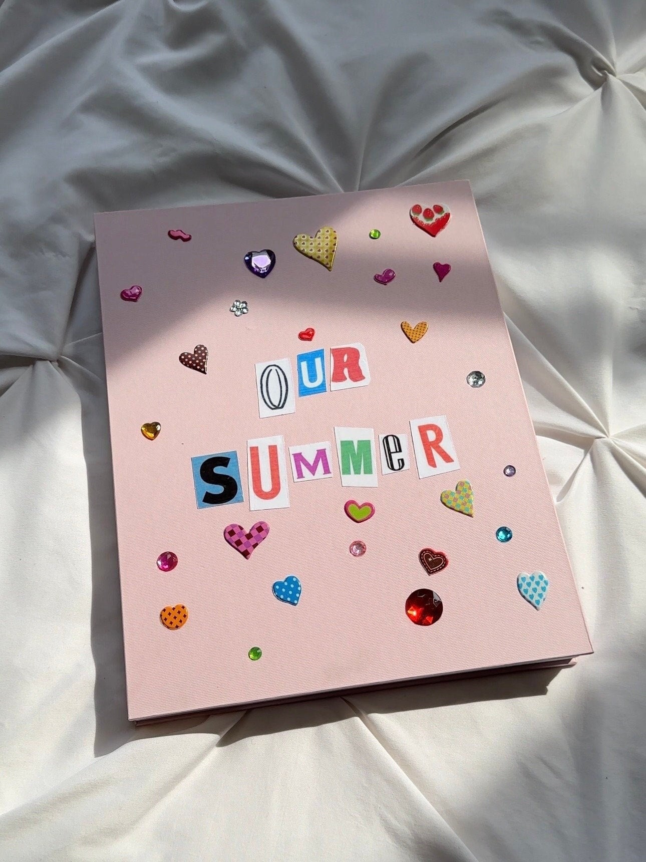 Light pink hardcover scrapbook with newspaper letters “Our Summer” on cover. Decorated with cute heart stickers and multicolored jewels.