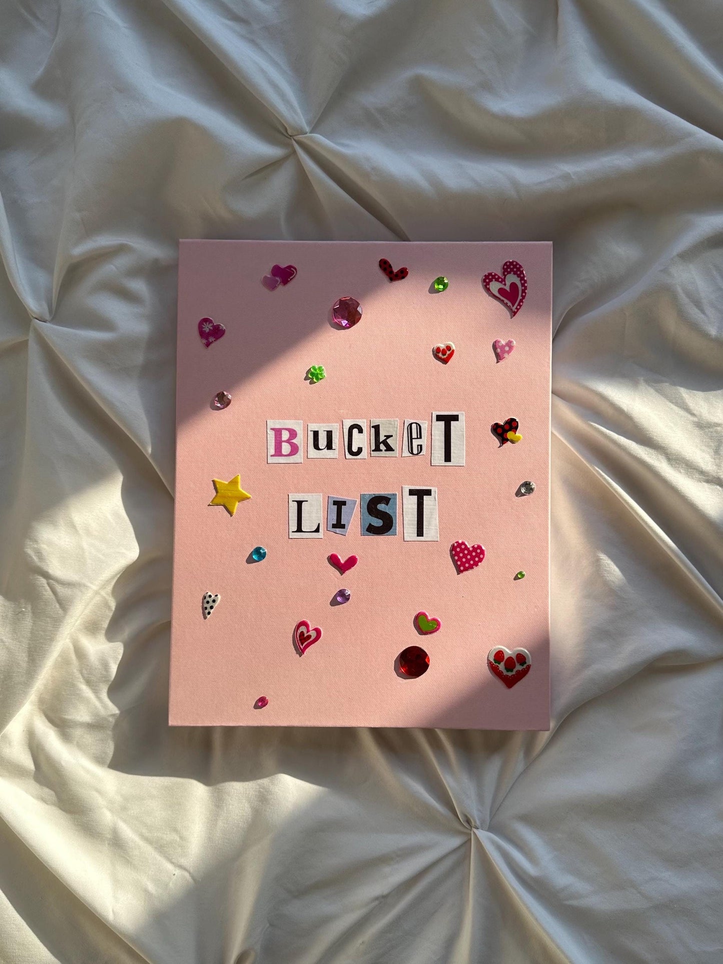 Light pink hardcover scrapbook with “Bucket List” written in newspaper letters. Decorated with heart and star stickers and jewels.