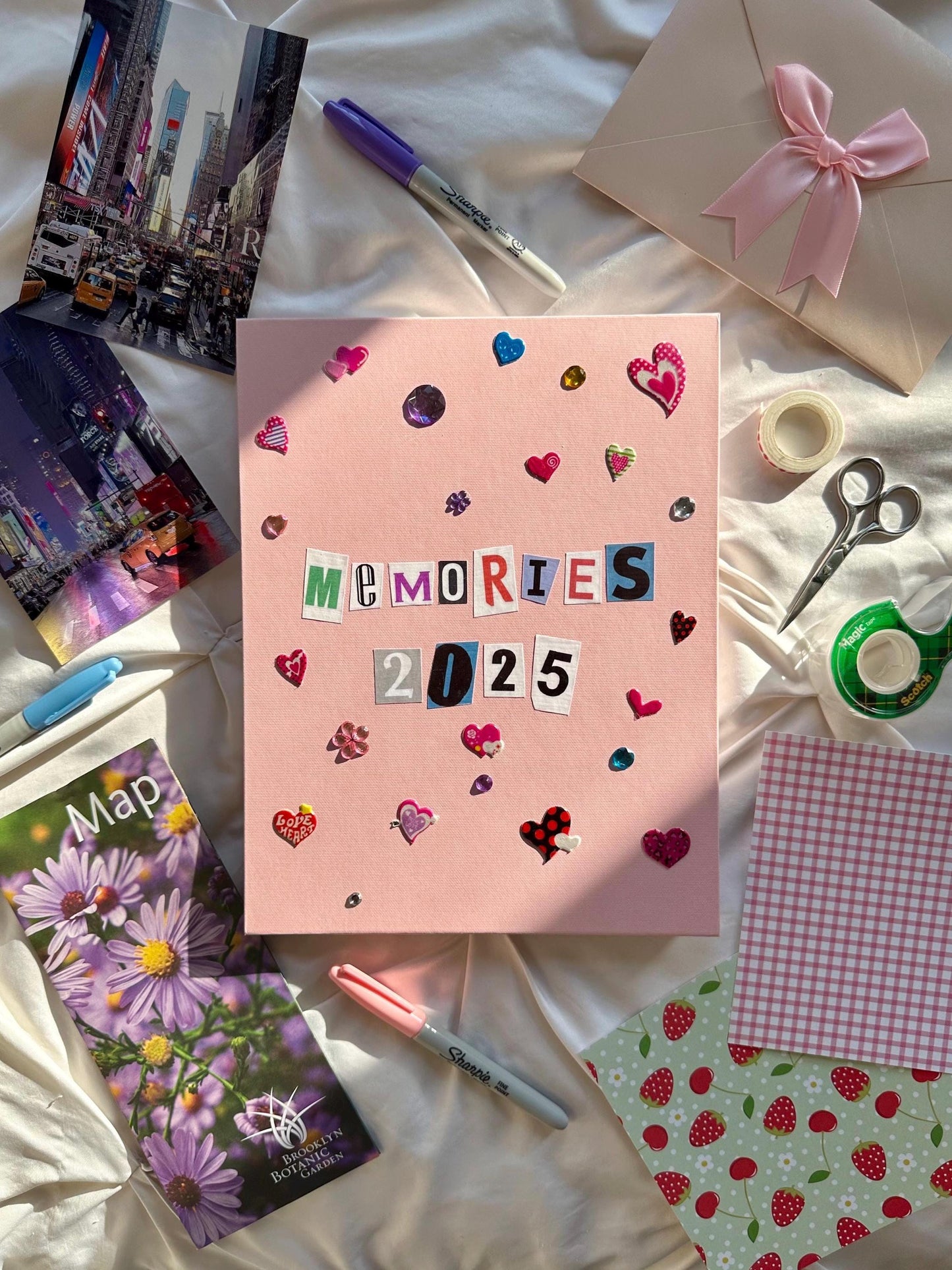 Light pink hardcover memory scrapbook for new years. Memories 2025 on cover in newspaper letters. Decorated with multicolored heart stickers and jewels.