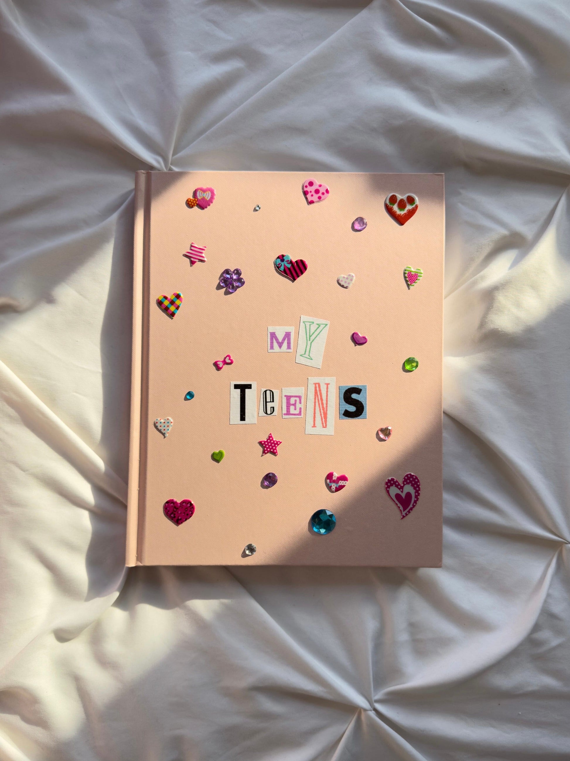 Light pink hardcover scrapbook with newspaper paper letters reading ‘My Teens’. Decorated with multicolored puffy heart stickers and jewels.