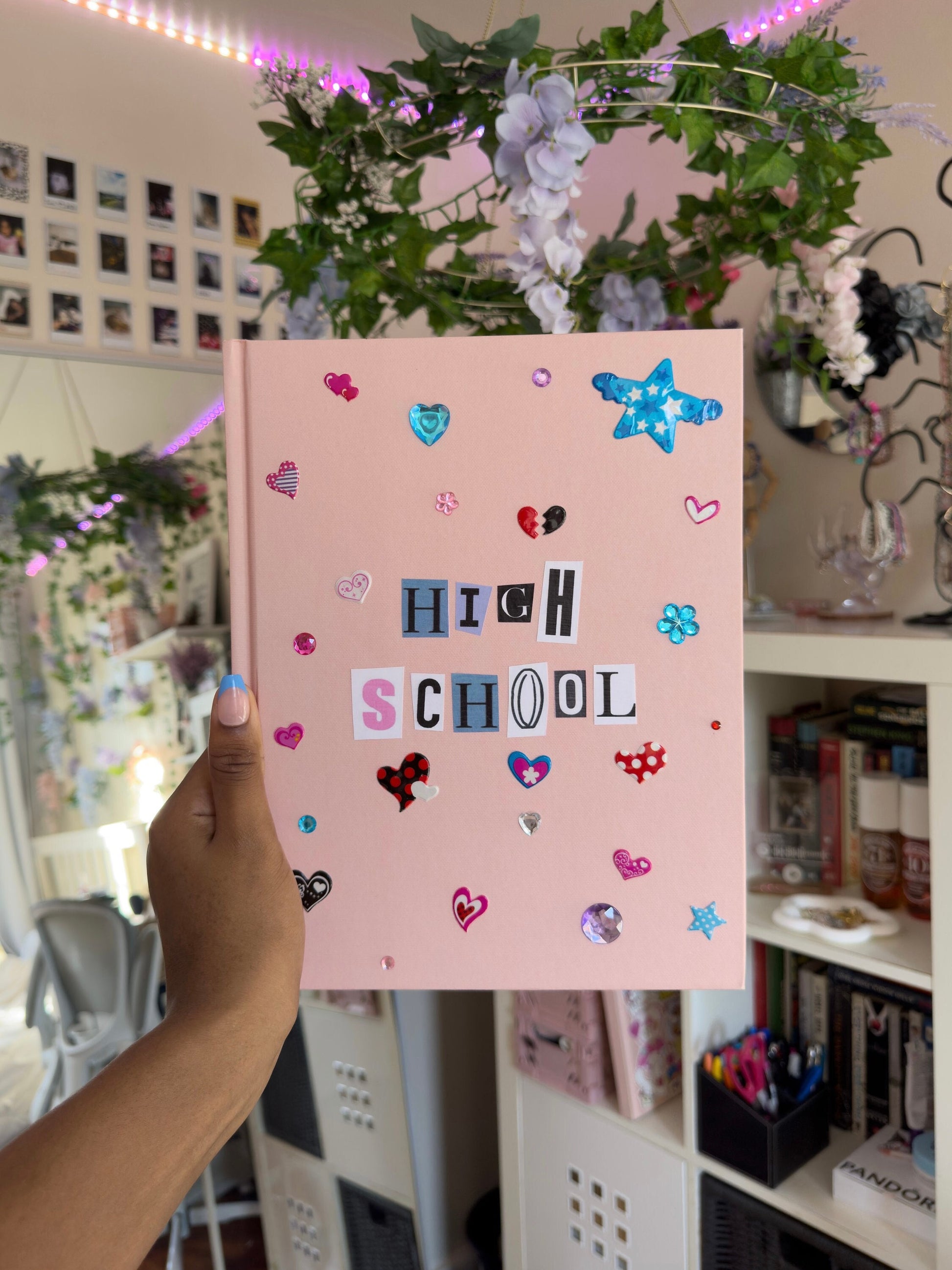 Large light pink hardcover scrapbook with newspaper letters reading “High School”. Decorated with cute heart and star stickers and jewels. Held up against an aesthetic girly bedroom background.