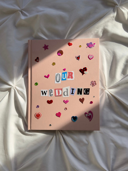 Light pink hardcover scrapbook with newspaper letters reading “Our Wedding”. Decorated with cute heart stickers and multi-colored jewels.