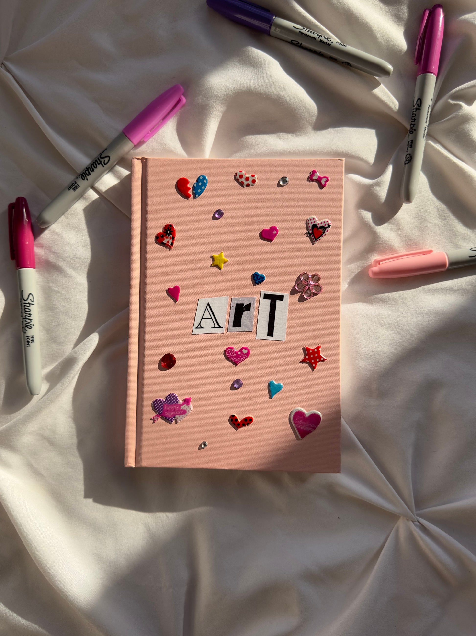 Light pink hardcover mini scrapbook with newspaper letters reading “Art”. Decorated with cute multicolored heart stickers and jewels.