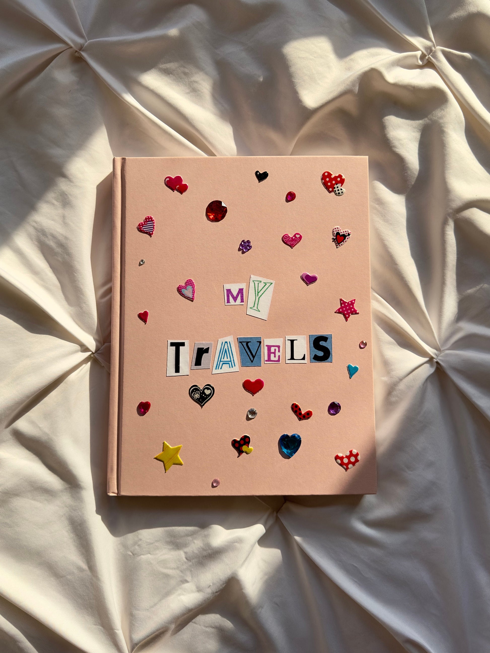 Light pink hardcover scrapbook with newspaper letters reading “My Travels”. Decorated with cute heart and star stickers and jewels.