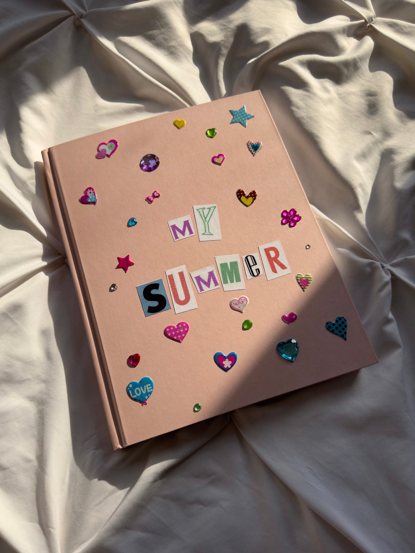 Light pink hardcover scrapbook lying in the sun. Decorated with newspaper letters reading “My Summer” and cute heart stickers and jewels.