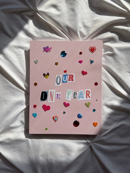 Light pink hardcover scrapbook with “Our One Year” the cover written in newspaper letters. Decorated with colorful heart stickers and jewels.