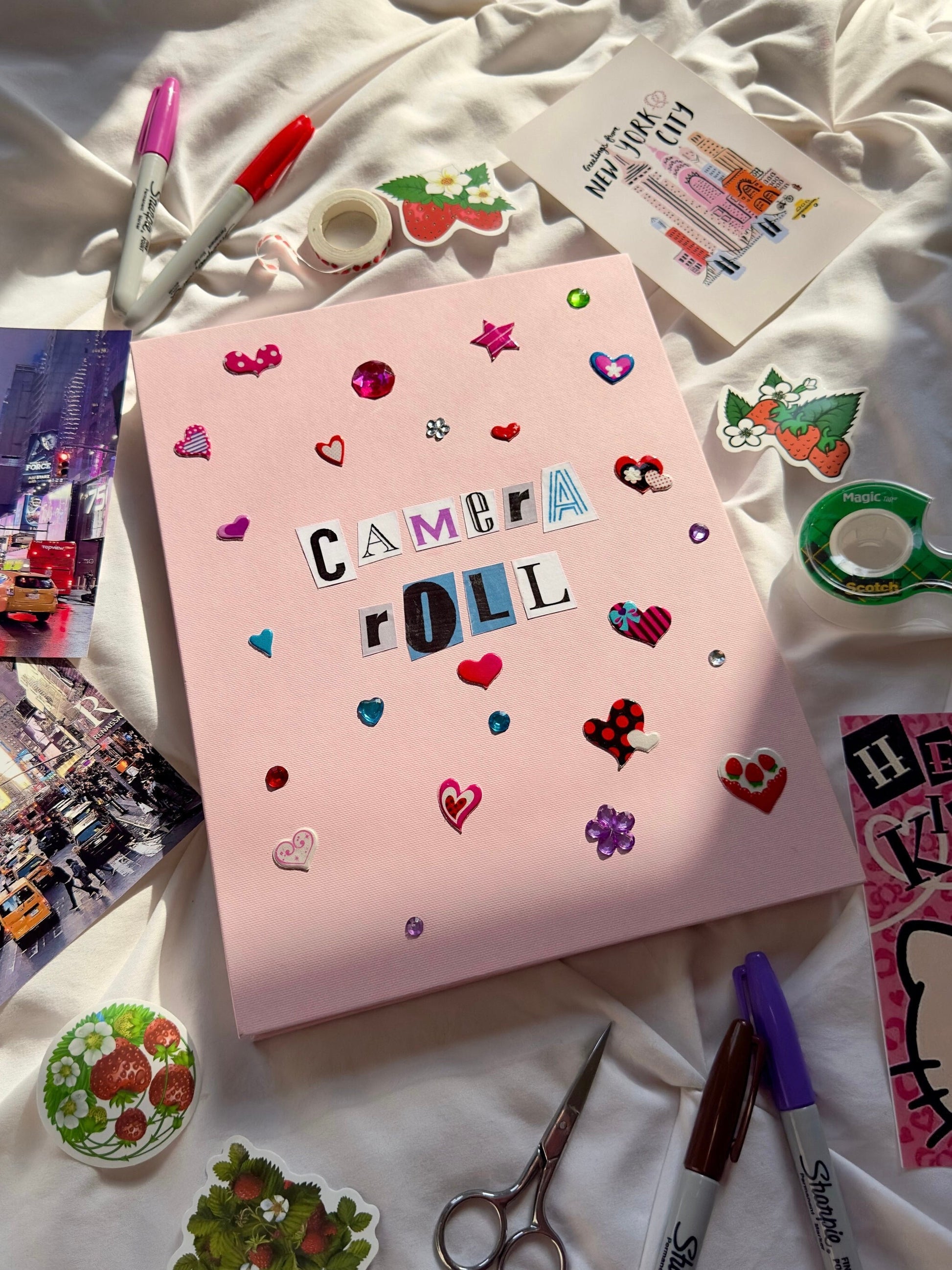 Light pink hardcover scrapbook with newspaper letters spelling “Camera Roll”. Decorated with cute heart stickers and jewels.