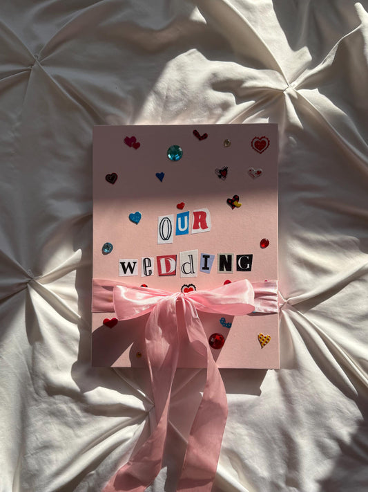 Light pink hardcover scrapbook with “Our Wedding” in newspaper letters on cover. Decorated with cute heart stickers and jewels. Pink bow wrapped around the scrapbook.